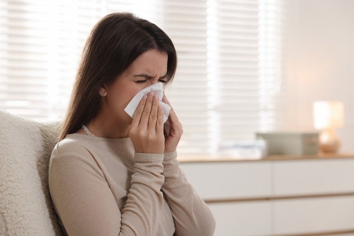 A woman sitting on a couch blowing her nose due to nasal congestion.