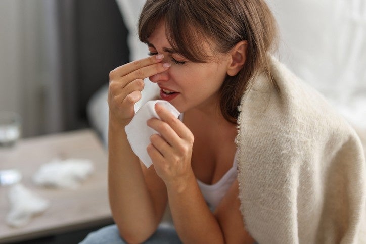 A woman seated on a bed holding a tissue and appearing unwell.